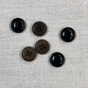 May include: Six buttons, four brown and two black, arranged on a light gray fabric background. The buttons have four holes each.