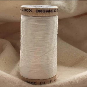 Puede incluir: Una bobina de hilo de algodón orgánico blanco con una etiqueta de madera que dice "100% Algodón Orgánico" y "Hecho en la UE."