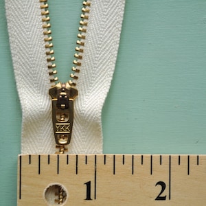 May include: A white zipper with a gold metal slider and pull tab. The zipper is measured against a wooden ruler with markings for 1, 2, and 3 inches.