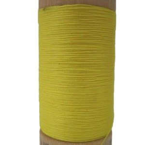 Puede incluir: Una bobina de hilo amarillo brillante con la etiqueta "100% Algodón Orgánico" y "100m / 110y".