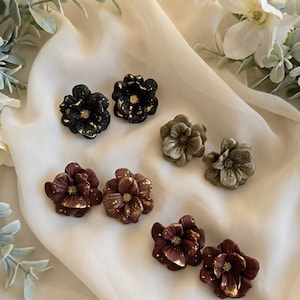 May include: An assortment of floral-shaped earrings in various colors, including black, burgundy, and olive green. Each pair features gold accents, creating a striking contrast. The earrings are displayed on a cream-colored fabric, showcasing their intricate details.