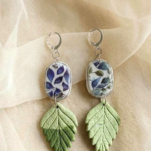 Pode incluir: Um par de brincos pendurados com um design de folha. Os brincos têm ganchos prateados e tops ovais transparentes com um padrão floral azul e branco. A parte inferior dos brincos são formas de folhas verdes.