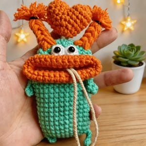 Puede incluir: Un monstruo de ganchillo hecho a mano en tonos turquesa y naranja. El monstruo tiene ojos grandes, una boca anaranjada ancha y pelo naranja. Un pequeño corazón naranja está en la parte superior de la cabeza. Una cuerda beige está unida al monstruo.