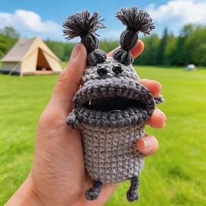 以下が含まれることがあります： グレーの色合いの手編みかぎ針編みのモンスター型ポーチ。ポーチは大きな開いた唇、2つの黒いビーズの目、2つの房状の髪のようなアクセントが特徴です。ポーチは、緑の草とテントを背景に、手で持たれています。