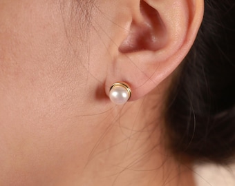 May include: A close-up of a pearl stud earring with a gold-colored setting. The earring is round and has a lustrous, white pearl. The gold setting encircles the pearl, creating a classic and elegant look.