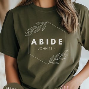 May include: Olive green t-shirt with the word "ABIDE" inside a white geometric design, with the text "JOHN 15:4" below. The shirt has a crew neck and short sleeves.