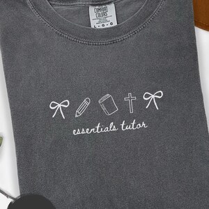 以下が含まれることがあります： 白の刺繍デザインが施されたペッパーカラーのTシャツ。デザインにはリボン、鉛筆、本、十字架、もう一つのリボンが含まれており、下に「essentials tutor」という言葉があります。シャツは折りたたまれています。