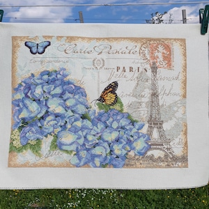 May include: Cross-stitch embroidery featuring a blue hydrangea and two butterflies against a vintage postcard design with the Eiffel Tower. The text "Carte Postale" is visible. The embroidery is displayed outdoors, hanging on a clothesline.