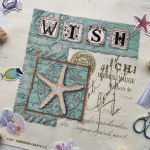 Può includere: Un disegno a punto croce con la parola "WISH" in blocchi decorativi, con una stella marina ed elementi in stile vintage. Adesivi di un granchio, pesce, conchiglie e un faro accompagnano il disegno. Il disegno è su un tessuto color crema.