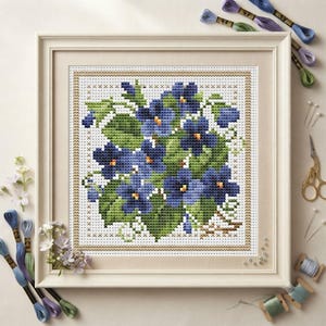 May include: A framed cross-stitch embroidery featuring a cluster of deep blue violet flowers with green leaves on a white grid background. The square piece is encased in a light-colored wooden frame, surrounded by embroidery floss and sewing tools.