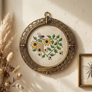 May include: A round, antique-style bronze frame featuring embossed bee and leaf motifs, enclosing a cross-stitch embroidery on light fabric. The design depicts a beehive, sunflowers, and bees. A small plaque at the bottom reads WILD-FLOWER MEADOWS.