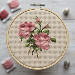 May include: A circular wooden embroidery hoop containing a piece of beige Aida fabric with a cross-stitch pattern of two blooming pink roses and a small rosebud, featuring green leaves and stems, secured by a metal tension screw at the top.