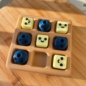 May include: A wooden tray with nine compartments, holding six blue and three yellow candies. The blue candies are spherical with black facial features. The yellow candies are square with black dots and a smiling face.