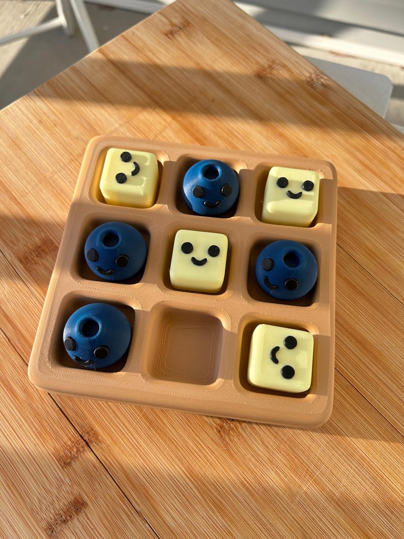 May include: A wooden tray with nine compartments, holding six blue and three yellow candies. The blue candies are spherical with black facial features. The yellow candies are square with black dots and a smiling face.