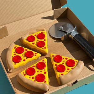Puede incluir: Cuatro juguetes con forma de porci&oacute;n de pizza con queso amarillo y pepperoni rojo est&aacute;n colocados en una caja de pizza de cart&oacute;n. Un cortador de pizza plateado con un mango negro tambi&eacute;n est&aacute; en la caja.
