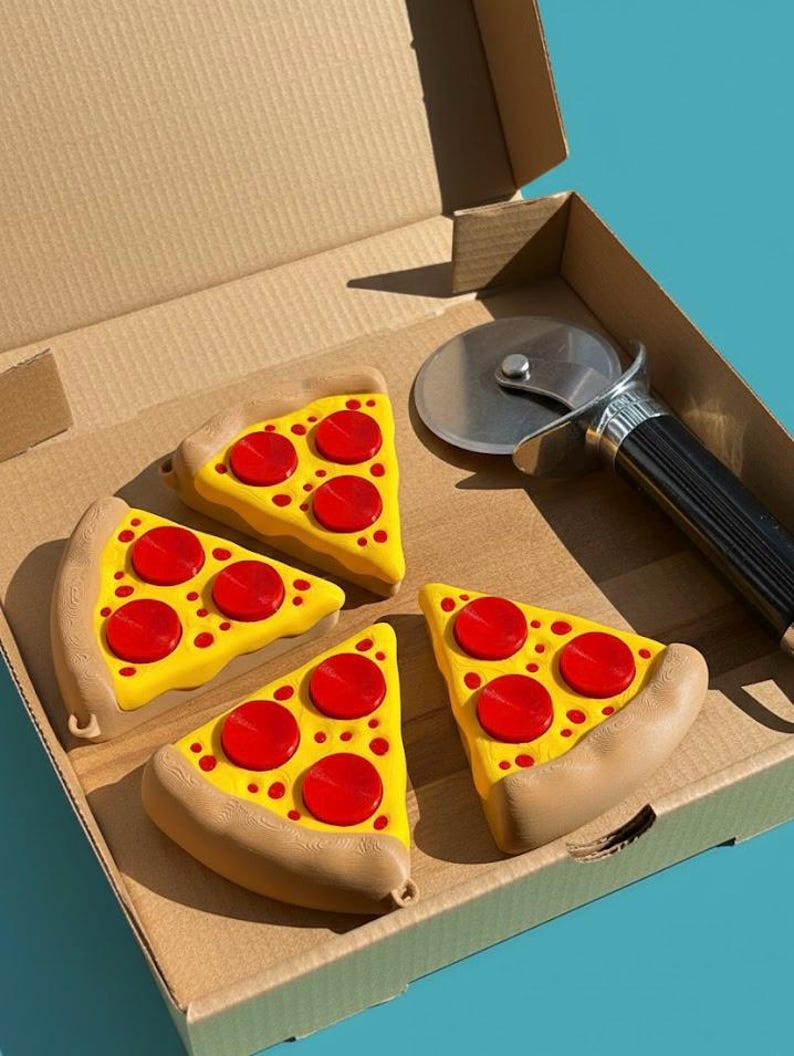 Puede incluir: Cuatro juguetes con forma de porci&oacute;n de pizza con queso amarillo y pepperoni rojo est&aacute;n colocados en una caja de pizza de cart&oacute;n. Un cortador de pizza plateado con un mango negro tambi&eacute;n est&aacute; en la caja.
