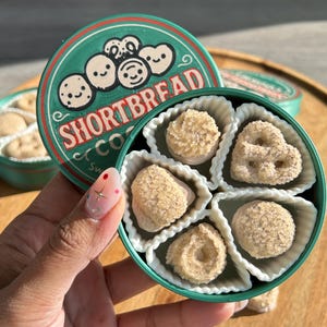 Könnte beinhalten: Eine grüne Dose Shortbread-Kekse mit einem Deckel mit einem Cartoon-Design und den Worten "Shortbread Co.". Die Dose ist geöffnet und zeigt verschiedene Keksformen in weißen Papierbechern. Die Kekse sind hellbraun.