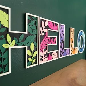 May include: A wall-mounted sign spelling the word HELLO in white-framed block letters. Each letter features a layered design with black backgrounds and various cutout leaf and foliage shapes in shades of lime green, dark green, pink, purple, orange, and blue.