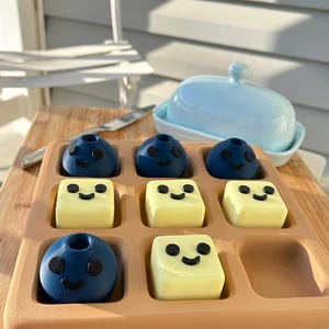 May include: A wooden tray holds nine food items: six are blue, spherical, and have black smiley faces, and three are yellow, square, and have black smiley faces. A light blue butter dish sits in the background. A butter knife rests on a wooden cutting board.