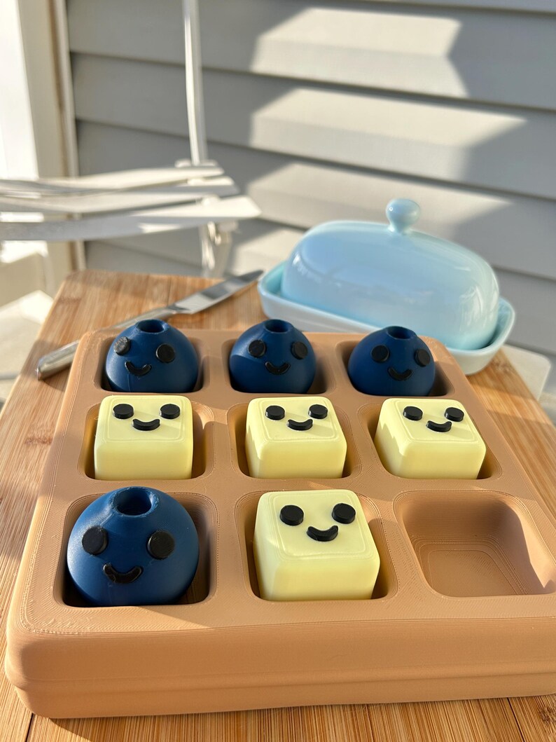 May include: A wooden tray holds nine food items: six are blue, spherical, and have black smiley faces, and three are yellow, square, and have black smiley faces. A light blue butter dish sits in the background. A butter knife rests on a wooden cutting board.