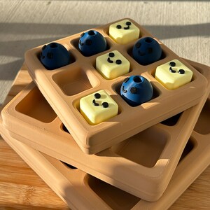 May include: A stack of three tan-colored waffle-shaped trays, each with a grid of square indentations. The top tray holds nine small, round, blue objects and yellow square objects, each with black dot details, resembling blueberries and butter.