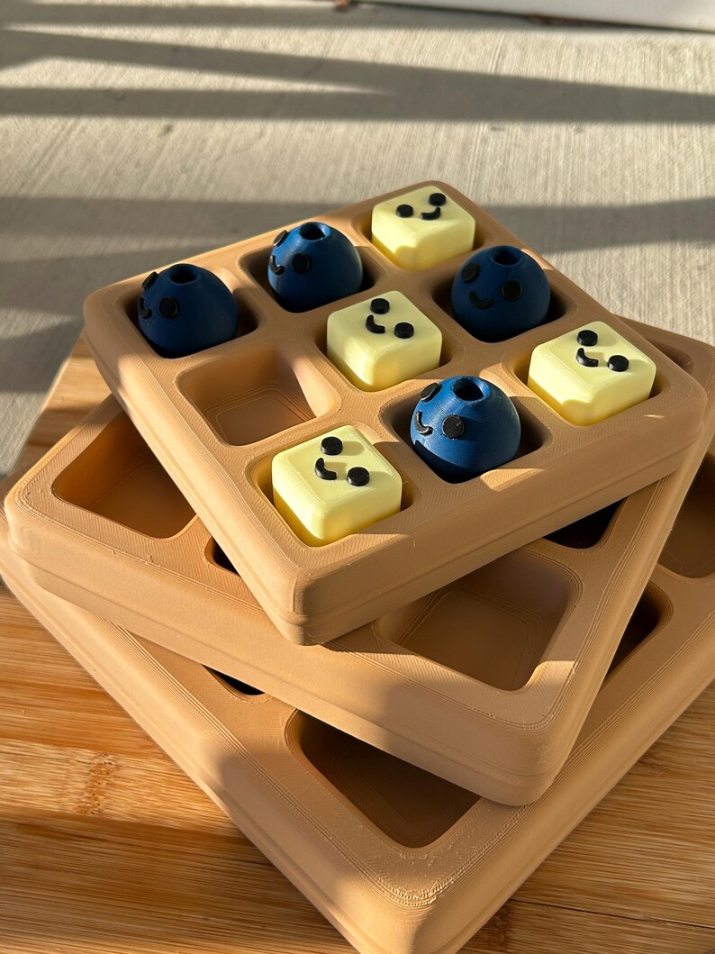 May include: A stack of three tan-colored waffle-shaped trays, each with a grid of square indentations. The top tray holds nine small, round, blue objects and yellow square objects, each with black dot details, resembling blueberries and butter.