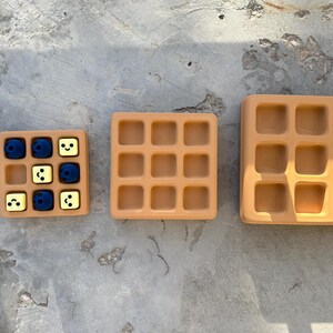 May include: Three tan-colored waffle-shaped trays of varying sizes. The smallest tray contains blue and yellow cubes. The trays are arranged on a gray concrete surface. The trays have a grid pattern.