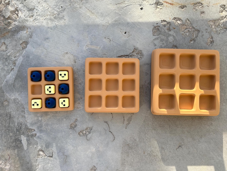 May include: Three tan-colored waffle-shaped trays of varying sizes. The smallest tray contains blue and yellow cubes. The trays are arranged on a gray concrete surface. The trays have a grid pattern.