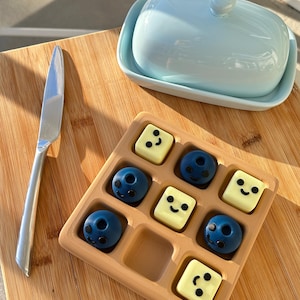 May include: A light blue ceramic butter dish with a lid and a silver butter knife rest on a wooden surface. A wooden tray holds nine chocolates, some round and some square, each with a smiling face.