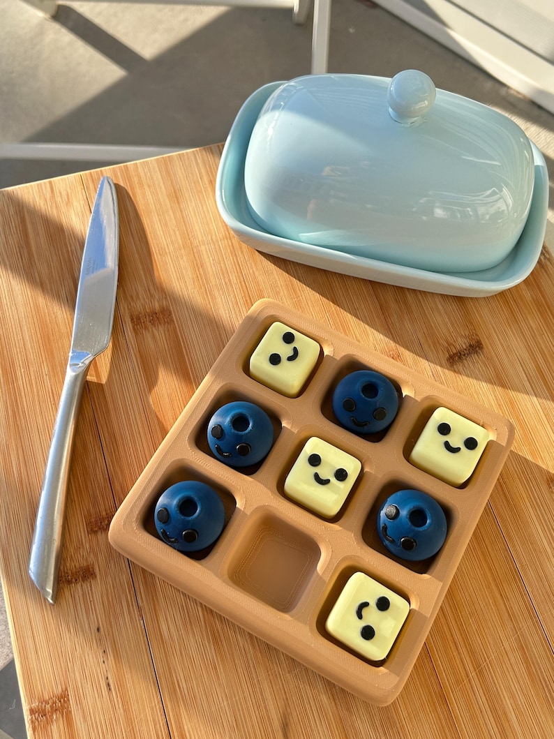 May include: A light blue ceramic butter dish with a lid and a silver butter knife rest on a wooden surface. A wooden tray holds nine chocolates, some round and some square, each with a smiling face.