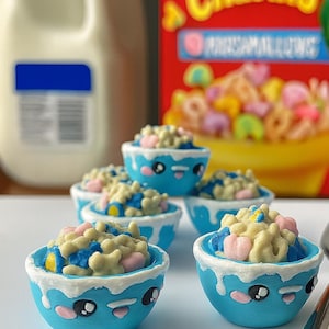 Puede incluir: Primer plano de varios cuencos azules pequeños llenos de cereales y malvaviscos. Los cuencos tienen bordes blancos y caras de dibujos animados. Una caja de cereales Lucky Charms y una jarra de leche están en el fondo.