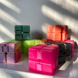 May include: A collection of colorful fidget cubes in various shades, including pink, green, orange, and black. The cubes are arranged on a white surface, with sunlight casting shadows. The cubes are designed with a modular, interlocking construction.
