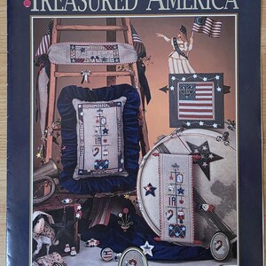 May include: A Treasured America craft pattern booklet featuring cross-stitch designs with patriotic themes. The cover displays various finished projects including a framed American flag, a tall vertical sampler, and small oval ornaments with stars.