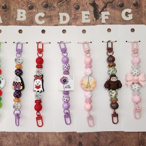 May include: A display of beaded keychains with various charms. The keychains feature a variety of colors, including green, red, purple, and pink. Charms include a Grinch face, a witch's hat, a ghost, a monkey, and a bow.