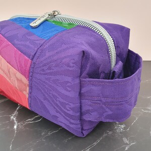 May include: A rectangular zipped pouch constructed from multi-coloured fabric panels in shades of purple, blue, green, pink, and red. The fabric features a subtle tonal jacquard pattern. A silver-toned metal zip runs along the top.
