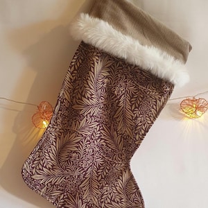 May include: A Christmas stocking featuring a burgundy and gold floral design. The top is brown corduroy with white faux fur trim. A cream ribbon is attached for hanging. Festive holiday decoration.