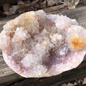 K&ouml;nnte beinhalten: Ein gro&szlig;er, klarer Quarzkristall-Cluster mit einem violetten Farbton und einigen orangefarbenen Kristallen. Der Cluster befindet sich auf einer Holzoberfl&auml;che.