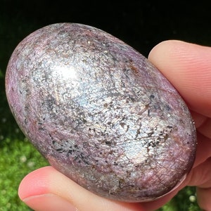 May include: A smooth, oval-shaped, polished gemstone with a silvery-purple hue and black flecks. The stone has a reflective surface and is likely a variety of ruby.
