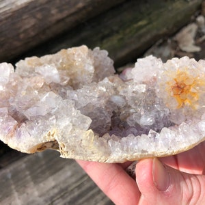 K&ouml;nnte beinhalten: Ein Amethyst-Cluster mit einer nat&uuml;rlichen, rauen Au&szlig;enseite. Die Kristalle sind hellviolett und haben eine durchscheinende Optik. Der Cluster hat einen Durchmesser von etwa 10 cm.
