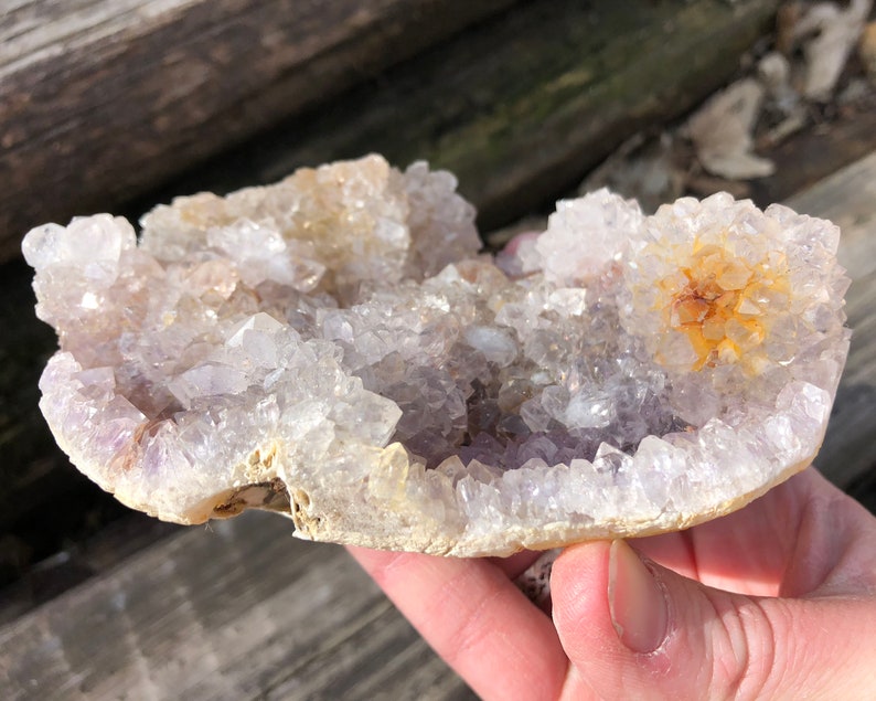 K&ouml;nnte beinhalten: Ein Amethyst-Cluster mit einer nat&uuml;rlichen, rauen Au&szlig;enseite. Die Kristalle sind hellviolett und haben eine durchscheinende Optik. Der Cluster hat einen Durchmesser von etwa 10 cm.