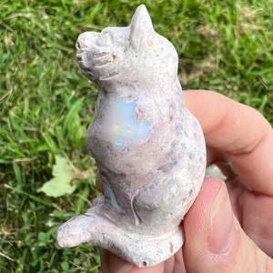 Pode incluir: Uma estatuinha de gato esculpida em pedra com uma superfície manchada de rosa e branco. O gato está sentado ereto com a cabeça inclinada para o lado. Um pedaço de opala iridescente está incrustado no peito do gato.