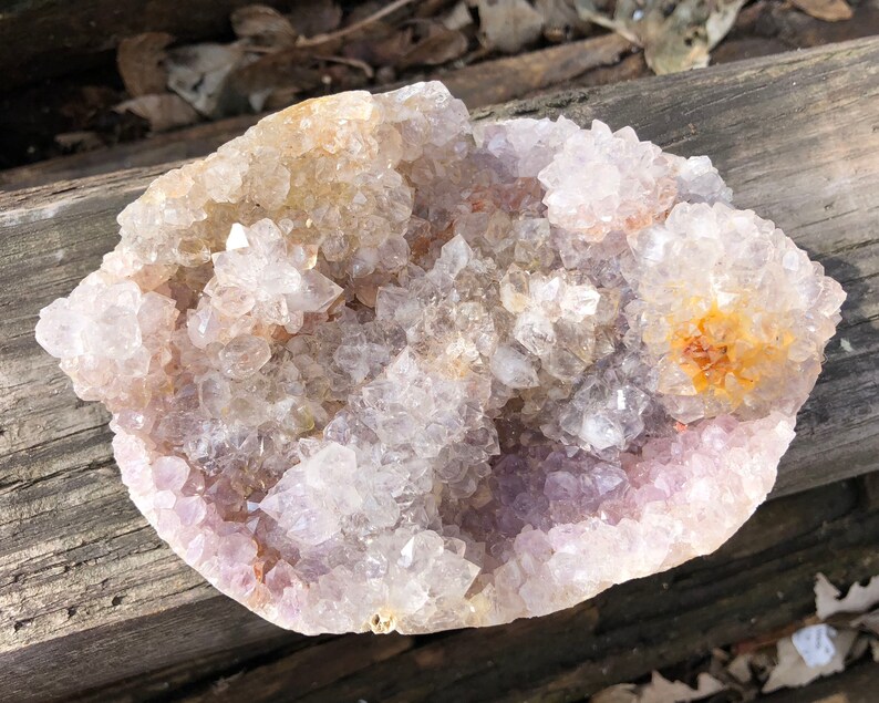 K&ouml;nnte beinhalten: Ein gro&szlig;er, klarer Quarzkristall-Cluster mit einem violetten Farbton und einigen orangefarbenen Kristallen. Der Cluster befindet sich auf einer Holzoberfl&auml;che.