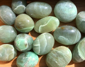 May include: A collection of smooth, green, polished stones with a cloudy, translucent appearance. The stones are arranged on a wooden surface.