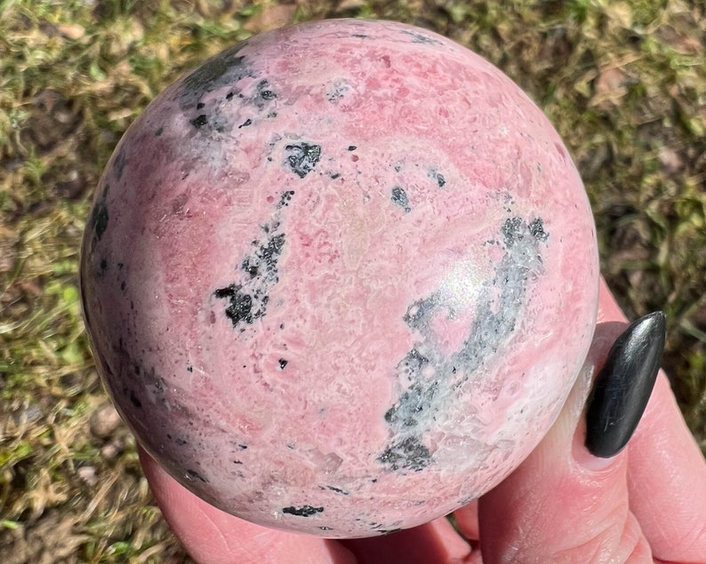 May include: A pink and black sphere of polished rhodochrosite, a type of crystal.