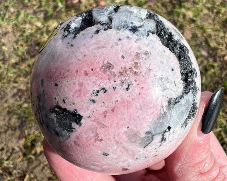 May include: Pink and black crystal sphere with a smooth, polished surface. The sphere has a unique pattern of black and white veins throughout the pink crystal.