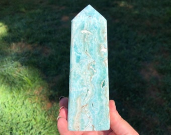 May include: A light blue, translucent, polished, aquamarine crystal tower with white and brown inclusions. The tower is a natural crystal formation and has a pointed top.