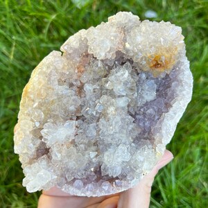 K&ouml;nnte beinhalten: Eine gro&szlig;e Amethyst-Geode mit einem Cluster aus klaren Quarzkristallen auf der Oberfl&auml;che. Die Geode ist hellviolett und hat eine nat&uuml;rliche, raue Textur.