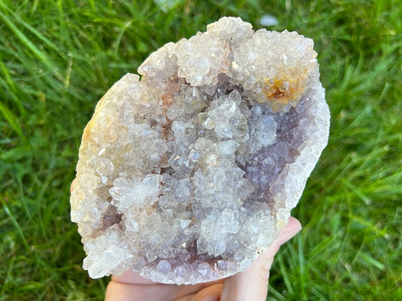 K&ouml;nnte beinhalten: Eine gro&szlig;e Amethyst-Geode mit einem Cluster aus klaren Quarzkristallen auf der Oberfl&auml;che. Die Geode ist hellviolett und hat eine nat&uuml;rliche, raue Textur.