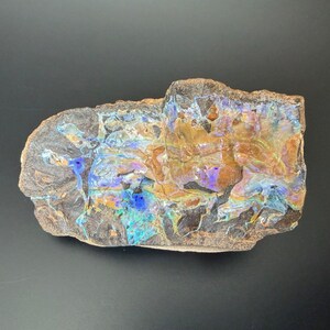 Puede incluir: Un espécimen de ópalo natural en bruto con una matriz oscura. El ópalo muestra colores vibrantes, incluyendo azules, verdes, naranjas y amarillos. La piedra tiene una forma irregular única, mostrando su estado bruto y sin pulir.