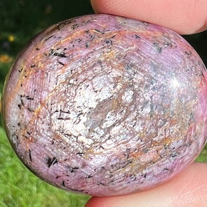 May include: A pink, purple and silver star ruby gemstone with black inclusions. The gemstone is polished and has a smooth, rounded shape.